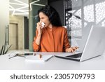 Young woman working sick at workplace inside office, worker sneezing has runny nose and allergy, hispanic sitting at table with laptop at work.