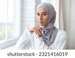 Close-up photo. Portrait of a young beautiful Muslim woman in a white hijab standing indoors by the window. He looks confidently and seriously into the camera.