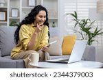 Small photo of Beautiful smiling woman studying online remotely sitting on couch in living room inside house, Hispanic woman using laptop to watch course video, writing thesis in notebook.