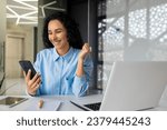 Happy and successful business woman reading message sitting at desk with laptop inside office, woman using app on smartphone, browsing online pages, and online video.