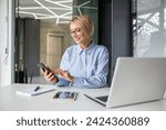Small photo of Careless attractive office worker using cellular phone while turning away from laptop in workplace. Inattentive blonde woman checking social media and mails instead of finishing business project.