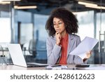 Small photo of Focused project manager with curly hair holding document and looking with hesitation at computer screen. Suspicious woman checking accuracy of printed report and comparing with electronic version.