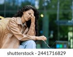 Small photo of Sorrowful woman with luxurious black curly hair holding pair of glasses while touching face in nasal area. Despondent female sitting on outdoors bench and resting after hard work day on fresh air.