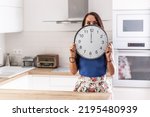 Small photo of Woman stands in the kitchen holding big clock showing exactly twelve o'clock.