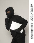 Small photo of Mysterious robber thief man wearing black hoodie and mask stealing laptop and sneak out. Isolated image on gray background