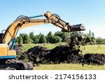 Small photo of An excavator with a wide bucket removes a layer of fertile soil. Preparatory work for digging a pit. Earth-moving equipment and earthworks