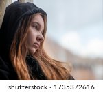 Low angle sad teen girl in black hoodie resting near window and looking depressed isolated teenager  maybe suffering from child abuse 