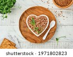 Heart-shaped bowl of boiled buckwheat porridge on old white wooden background. Gluten free ancient grain for healthy diet. Top view. Copy space.