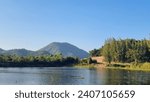 Small photo of Huai Tha koei reservoir at Ratchaburi city, Thailand. scenic view of water reservoir shows lake with pine forest at hill and bright blue sky. Huai Tha koei reservoir is a camp site destination.