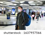 Small photo of Young European man in gray coat, protective disposable medical mask in airport. Afraid of dangerous N-CoV 2019 influenza coronavirus mutated and spreading in China. Blue backpack, suitcase on wheels.