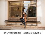 Handsome man holding takeaway coffee cup and mobile phone in hand. City life coffee and breakfast cafe in background. Man in brown coat Modern businessman
