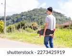 Skateboarder with his back to the camera holding a longboard on the street, with green hills in the background.