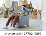 Young bearded man sitting on armchair and using laptop computer he moving to new apartment