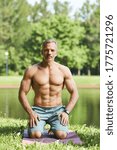 Small photo of Portrait of confident muscle-bound man with gray hair sitting on exercise mat in park with lake