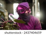 Small photo of Medium closeup of unrecognizable young woman wearing kerchief mask and goggles holding lamp examining plant leaves