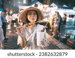 Poeple travel and eating street food concept. Happy young adult asian foodie woman holding bbq grilled skewers at outdoor vendor night market.
