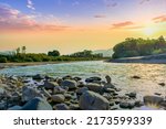 beautiful landscape of amazing sunset on a rough stormy mountain river with stones on foreground and green bushes and trees on sides