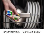 Close-up of young woman hand holding two colored capsule for the dishwasher. In the background, out of focus, is a dishwasher with clean dishes. Small depth of field.