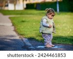 Small photo of Rascal little female child with braids jumping in a muddy puddle on a sunny spring day. Scoundrel 2 year old girl hopping in a small water plash by the grass. Kids exploration and freedom concept