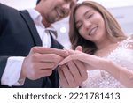 Small photo of Asian couple smile while gloom put diamond ring to bride's ring finger softly in their wedding ceremony