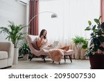 Young beautiful woman drinking rose wine and using laptop while sitting on comfortable pink armchair in home interior near window. Relax woman watching movie on laptop or make online purchases.