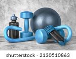Set of fitness equipment on a gray concrete background, front view, close-up. Dumbbells, sports bottle of water, jump rope, s-shaped leg exercise machine, fitness ball. Home workout.