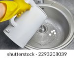 Small photo of A person in yellow rubber gloves pours hot water with steam from an electric kettle into a metal sink. Cleaning and disinfection of sanitary ware in the kitchen