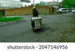 Small photo of The old man collects scrap metal on the streets of the city. Social problems of the elderly population of the city. Joyless unemployed man in age with financial problems is dragging an old refrigerato