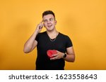 Handsome caucasian young man wearing black shirt isolated on orange background in studio keeping pink box, pointing finger on pulse, smiling. People sincere emotions, lifestyle concept.