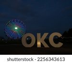 Small photo of Oklahoma City, OK USA - November 3, 2022: OKC sign made by Hugh Mead, photographed at the blue hour with the lit, stationary Wheeler Ferris Wheel in the background