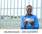 Small photo of A cheerful young man in a blue shirt is happily counting a stack of cash bills in front of a tall metal fence, seemingly outdoors during the daytime.