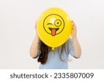A child girl hides his face behind a yellow winking emoticon on white background. Funny face. World Emoji Day