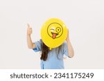 A child girl hides his face behind a yellow winking emoticon on white background. Funny face. World Emoji Day