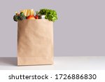 Small photo of Various grocery items in paper bag on white table opposite gray wall. Bag of food with fresh vegetables, fruits, pasta and canned goods. Food delivery, shopping or donation concept. Copy space.