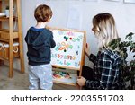 Kindergartener stand with back and learn english letters, alphabet and numbers on magnetic board. Educator sit near boy in playroom and check exercise. Reading and counting, study in daycare centre. 