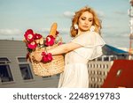 Small photo of Portrait of young beauteous woman with long wavy fair hair wearing white light summer dress, floral earrings, standing, holding straw bag with bouquet of pink, yellow flowers tulips, baguette bread.