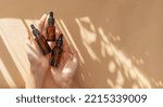 Small photo of Woman's hand holds three amber glass vials with pipette on beige background in rays of sunlight. Top view of unmarked containers with essential oil, moisturizing serum. Cosmetic product for body care.