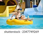 Boy and girl have fun on water slide in outdoor aquapark. Little children floating on yellow inflatable raft.