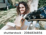 Small photo of Young long hair hippie man smiling playing guitar in park while resting against a tree. Hobby art activity for young adults. Authentic manhood moment.