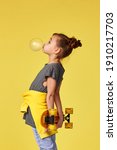 Small photo of funny little child girl with yellow skateboard blowing a big bubble gum over yellow background