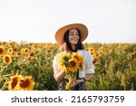 A happy little girl with freckles in a white shirt, skirt and straw hat holds a bouquet of sunflowers in a field of sunflowers.