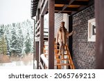 Beautiful caucasian woman walking down on wooden stairs of modern cottage and looking on snowy nature around. Winter holidays among mountains. Lifestyles concept.