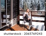 Relaxed woman sitting on wooden terrace with cup of fresh coffee or tea and enjoying morning among snowy forest. Winter holidays among peaceful nature.