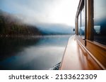 Close up of modern wooden boat floating on lake Konigsee in Berchtesgaden. Winter fog over forest and mountains with reflection on water surface.