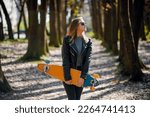 Woman standing at forest alley and carrying longboard at summer time. Skater in casual wear hold longboard in hands 