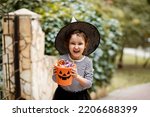 Small photo of Little cute girl in witch costume holding jack-o-lantern pumpkin bucket with candies and sweets. Kid trick or treating in Halloween holiday