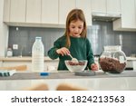 A cute girl prepearing quick breakfast made of chocolate cereal balls and milk stirring with a spoon in a huge light kitchen at home