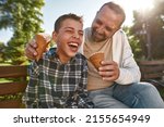 Cheerful caucasian father and teenage son with cerebral palsy eat ice cream and joke on bench in sunny park. Family relationship and enjoy time together. Disability care, treatment and rehabilitation