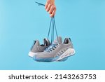 Hand holds hanging gray running sneakers by the laces on blue pastel background.  Hand with a new sport shoe. Stability and cushion running shoes. Close up.