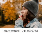 Small photo of Young woman outside autumn park fall and smoking tobacco device electronic cigarette heater. Smoke and steam system with sticks inside, image with copy space. Harmful habit harm to health lungs